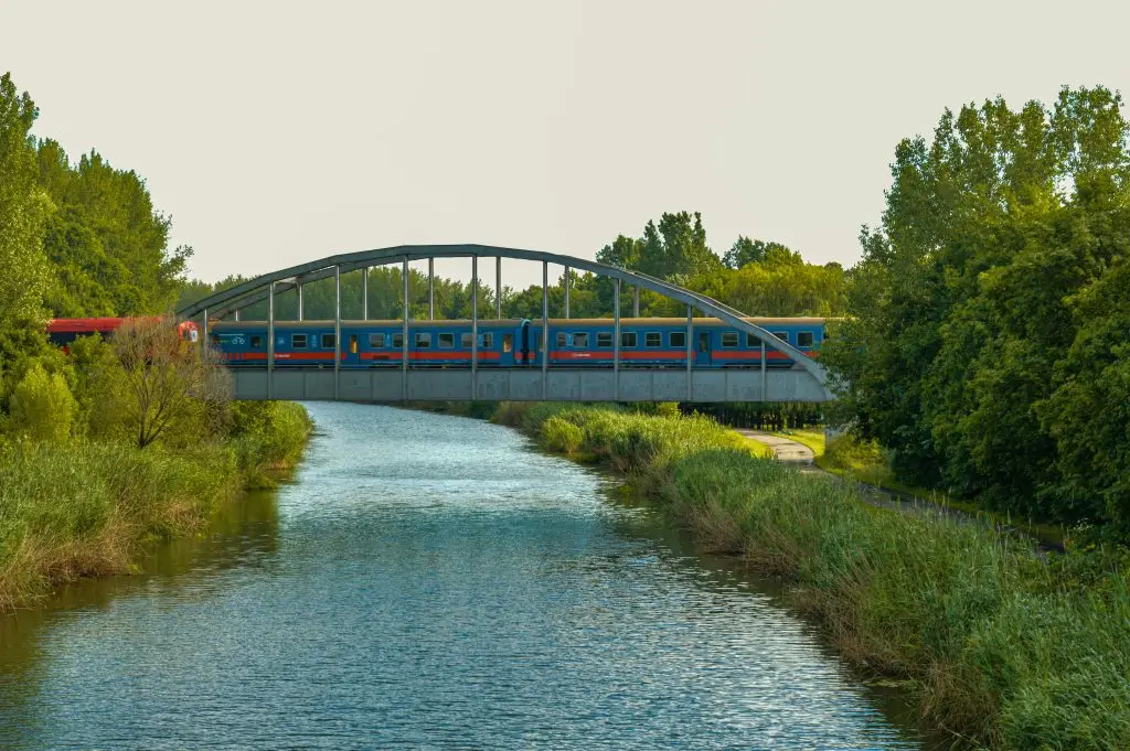A vibrant train crosses a bridge over a tranquil river surrounded by lush greenery.